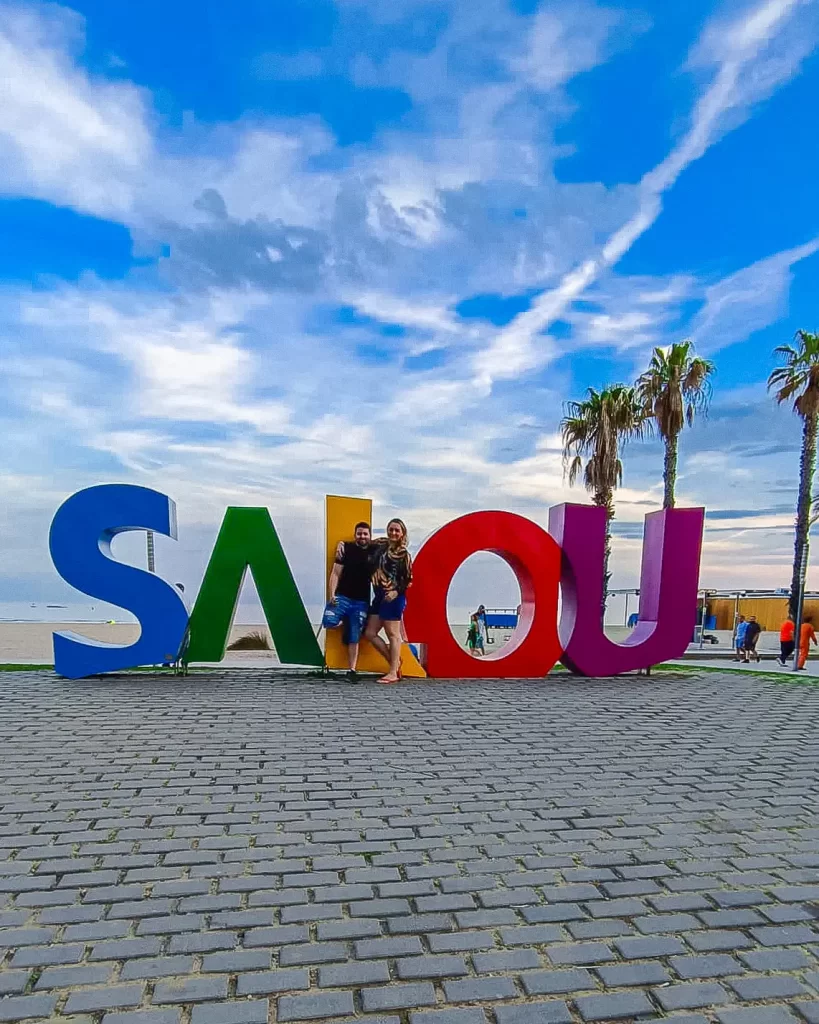 Casal posando em frente ao letreiro colorido de "SALOU" com palmeiras e o céu azul ao fundo.