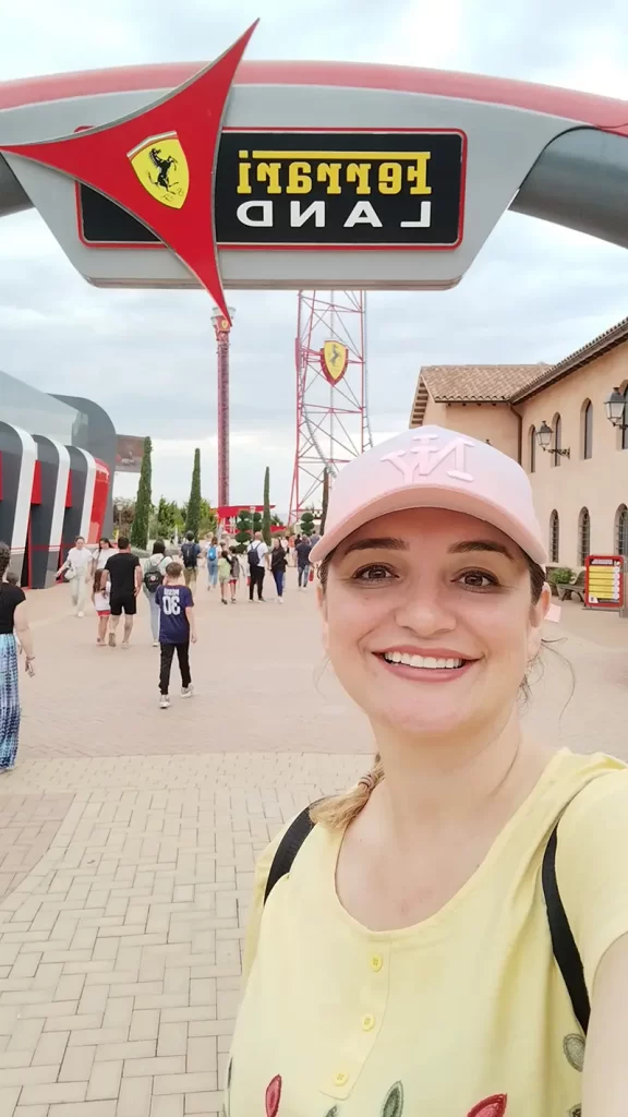 Camila sorrindo com um boné rosa na entrada do Ferrari Land em Salou, com o logo da Ferrari e uma montanha-russa ao fundo.