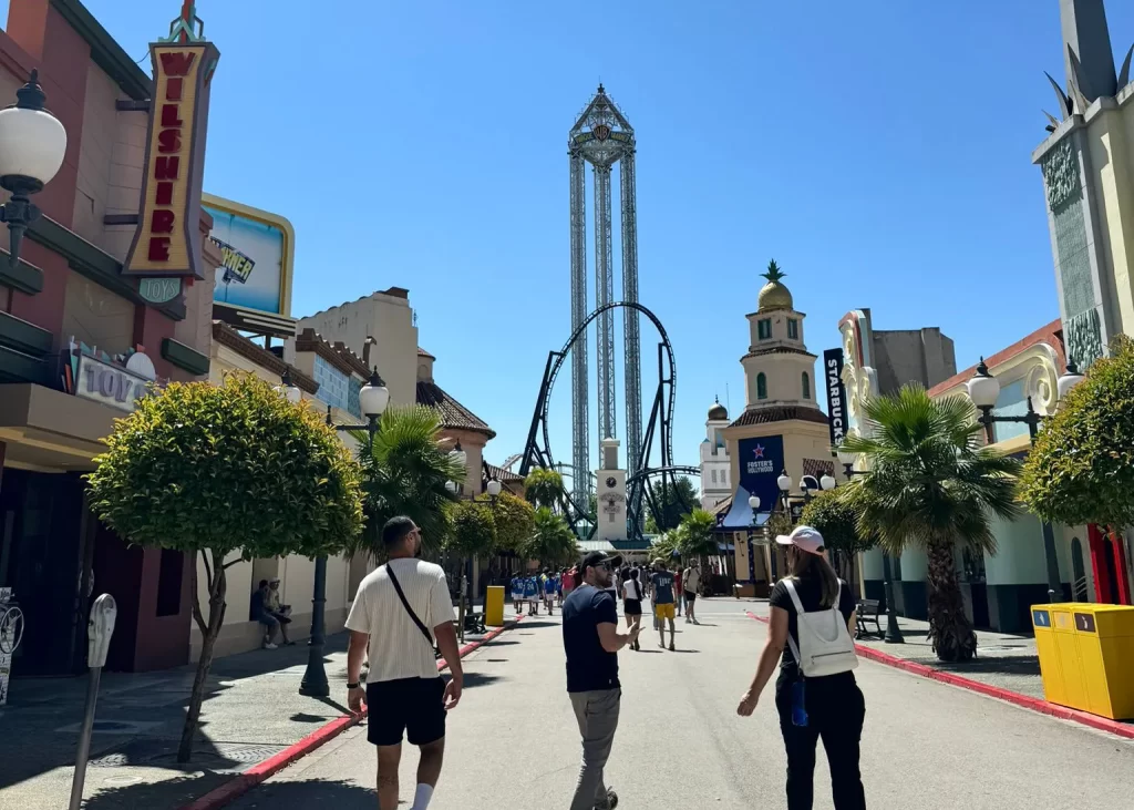 Vista da entrada principal do Parque Warner Madrid em um dia ensolarado, com pessoas caminhando em direção às atrações. No centro, destaca-se uma torre alta de queda livre e uma montanha-russa com loops e curvas.