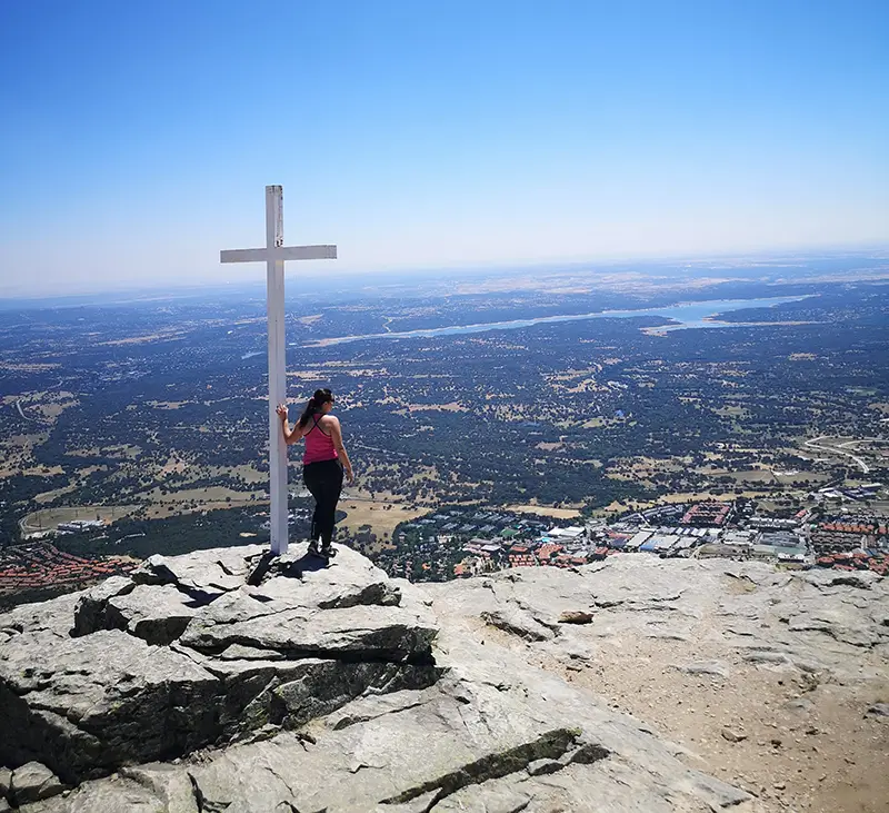 Mulher de camiseta rosa e calça preta tocando uma cruz branca no Pico de Abantos, com vista panorâmica de San Lorenzo de El Escorial e paisagens naturais ao fundo em um dia claro e ensolarado.