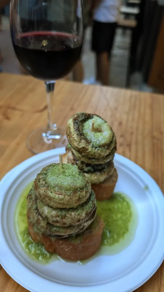 Um prato de tapas com cogumelos grelhados empilhados sobre uma fatia de pão, acompanhado por um copo de vinho tinto em uma mesa de madeira.