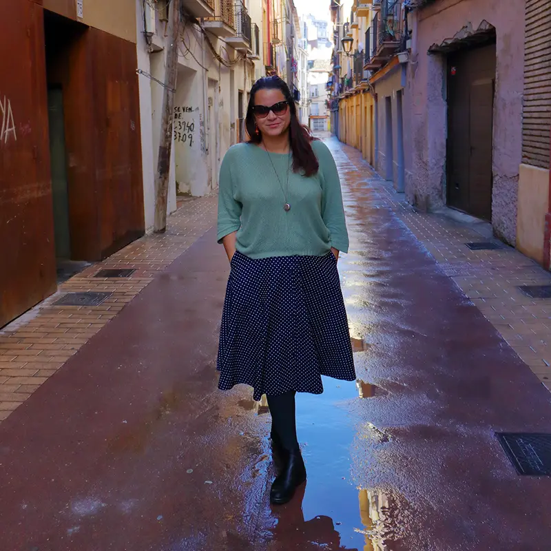 Eu caminhando por uma rua estreita e charmosa em Zaragoza, Espanha, com edifícios históricos de ambos os lados e o chão molhado refletindo a luz suave do dia.