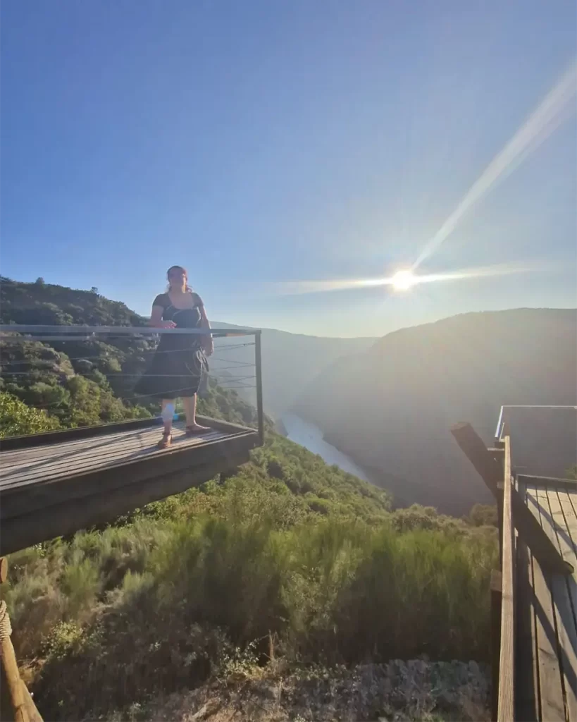 Pessoa de pé em uma das plataformas de madeira do Mirador, com vista para o rio e montanhas ao fundo, durante o pôr do sol.