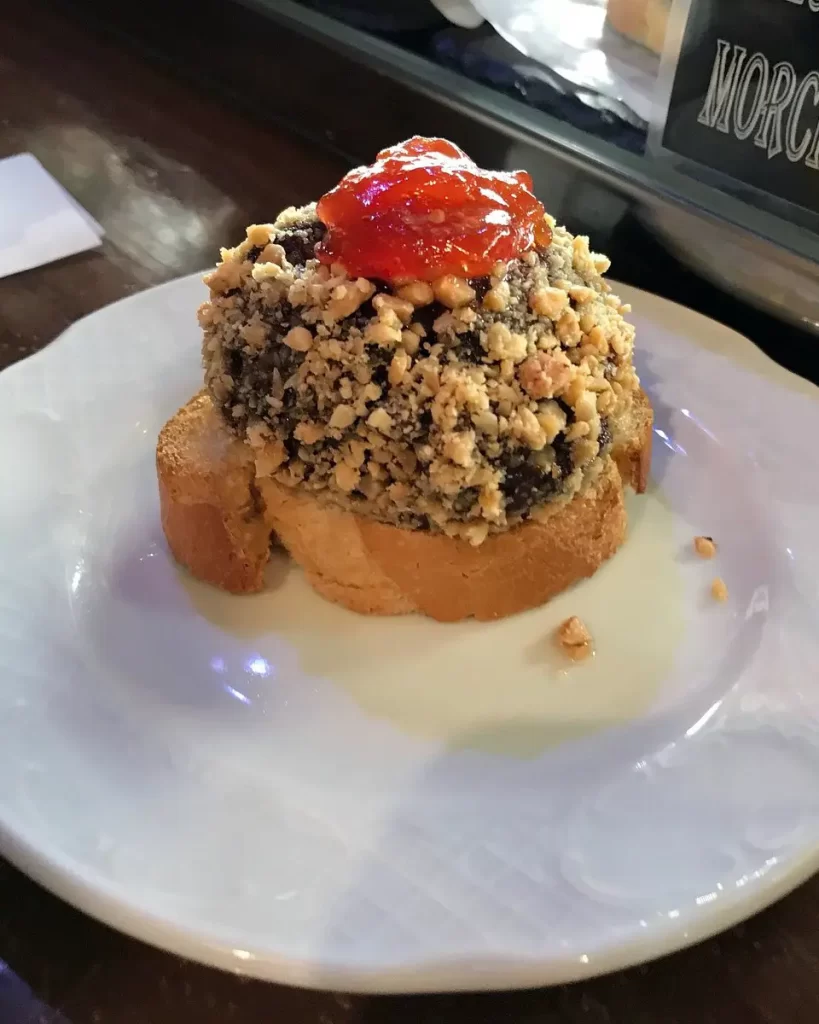 Tapa de morcilla servida no Bar Victorinos, coberta com uma crosta de nozes e uma porção de geleia vermelha no topo, sobre uma fatia de pão.