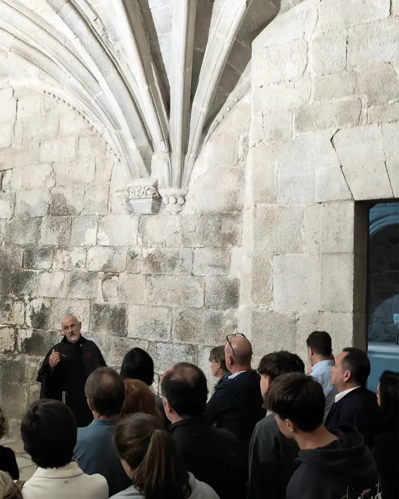 Ator vestido de monge conduzindo uma visita teatralizada no Monasterio de Santo Estevo, enquanto um grupo de visitantes o ouve atentamente em um salão de pedra.