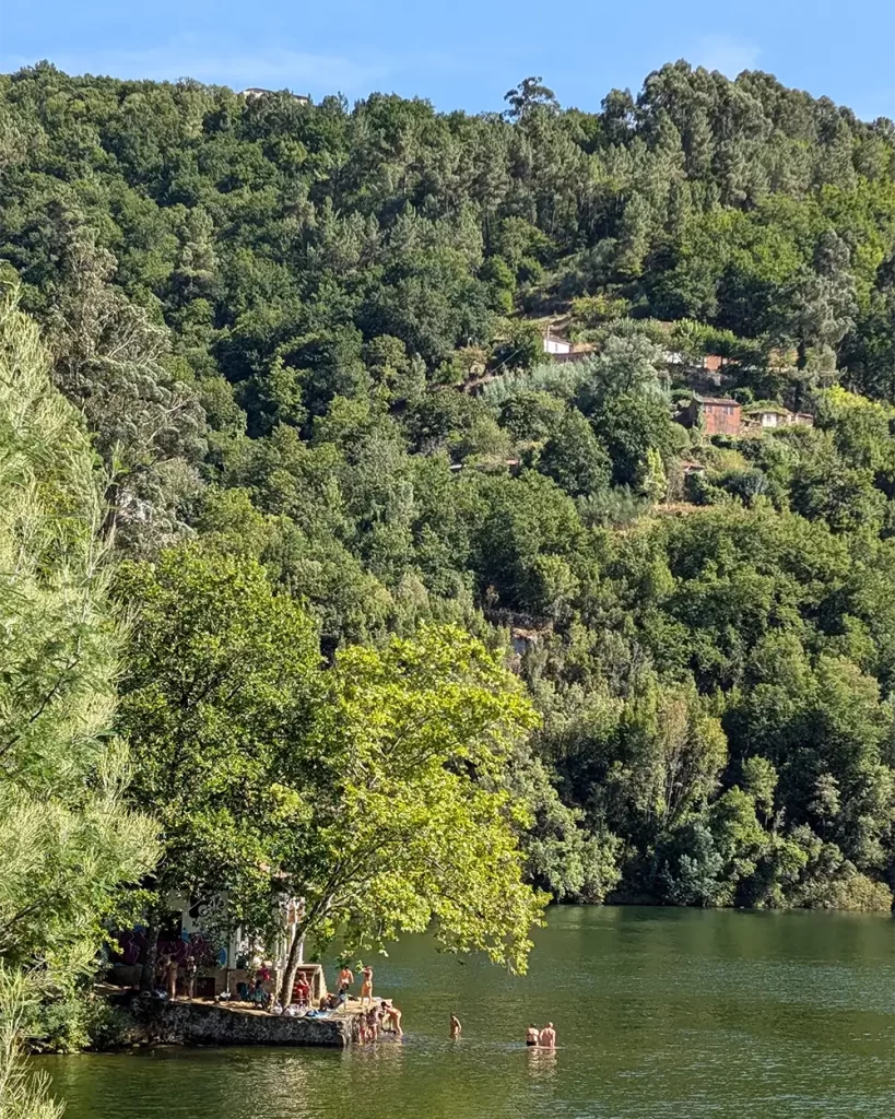 Vista de uma pequena construção à beira do Rio Miño, cercada por árvores e pessoas nadando. 