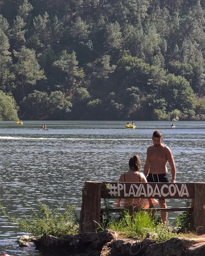 Casal sentado em um banco com a placa “#PlayaDaCova” à beira do Rio Miño, observando as águas calmas.