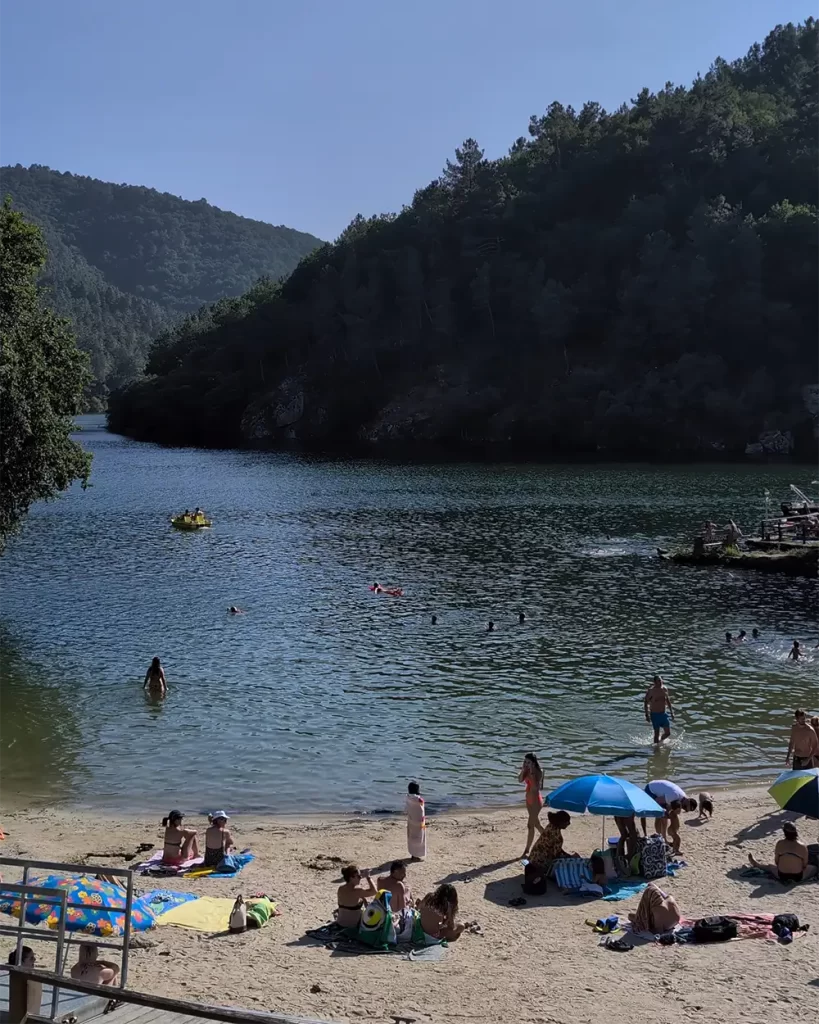 Pessoas curtindo um dia ensolarado na Praia Fluvial de A Cova, com montanhas verdes ao fundo.