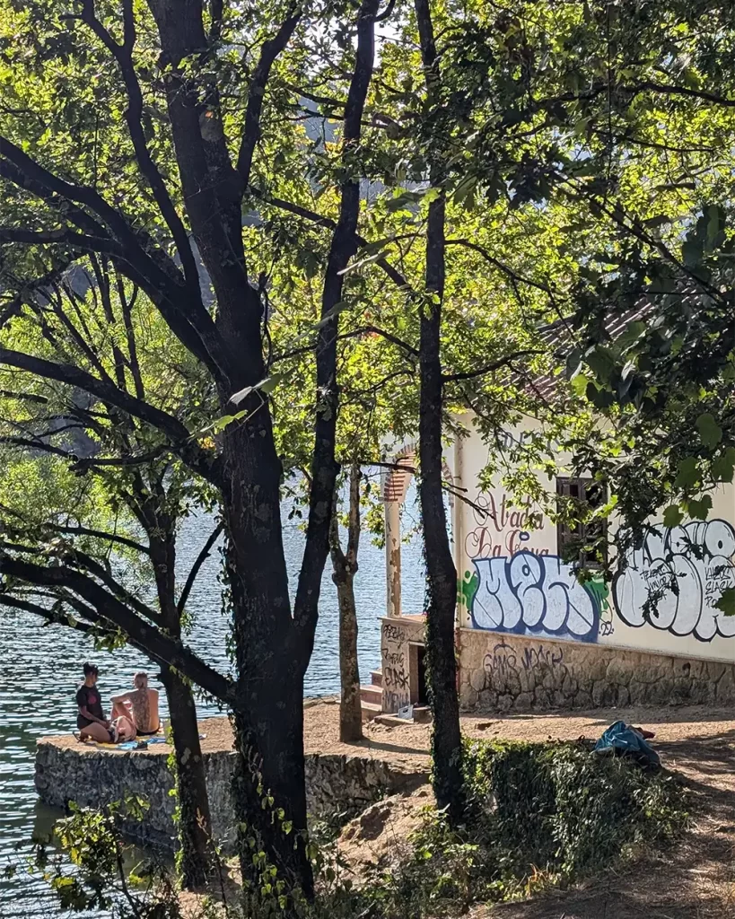 Pessoas sentadas à beira do Rio Miño, parcialmente escondidas entre as árvores, perto de uma construção com grafites na Praia Fluvial de A Cova.