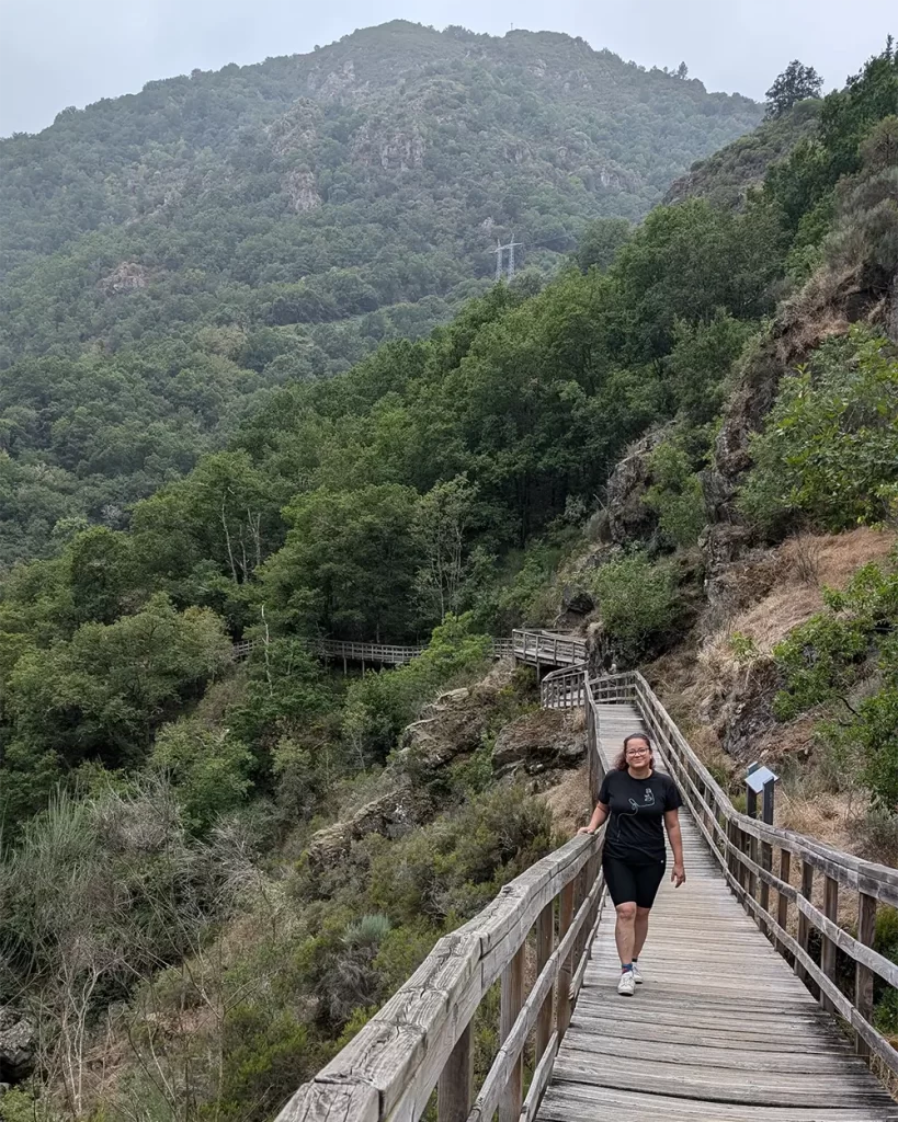 essoa caminhando sobre passarelas de madeira na Trilha do Rio Mao, com montanhas e vegetação densa ao fundo.