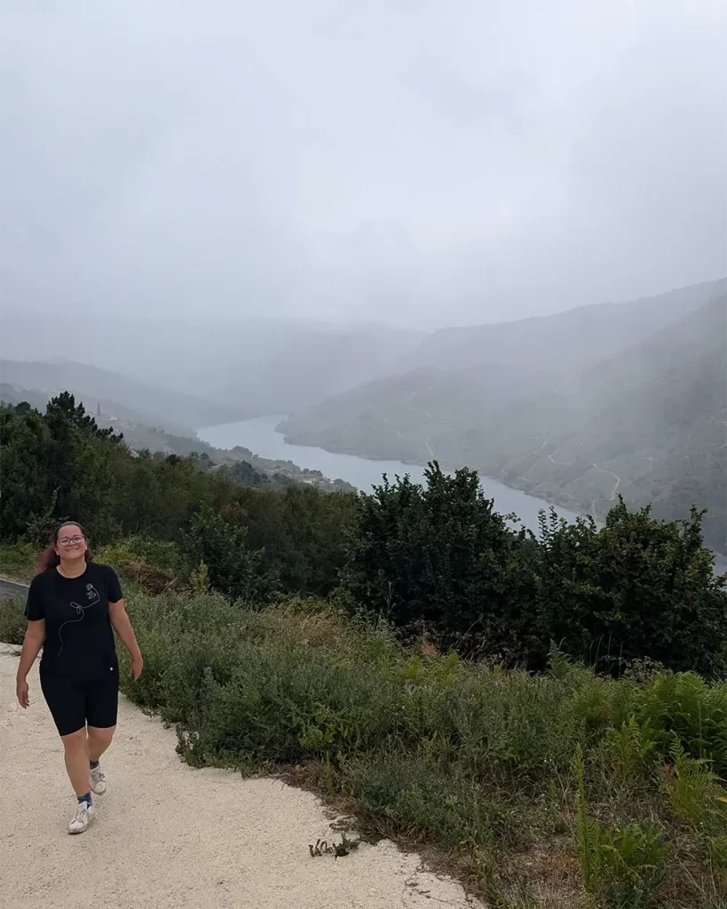 Pessoa caminhando em uma trilha de cascalho com o Rio Sil e montanhas cobertas por neblina ao fundo.