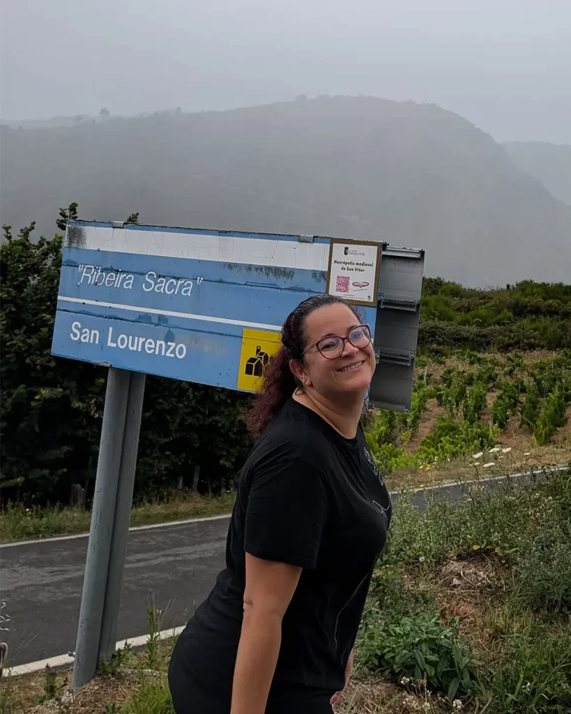 Pessoa sorridente posando ao lado de uma placa de sinalização que indica "Ribeira Sacra" e "San Lourenzo", com uma paisagem montanhosa ao fundo.