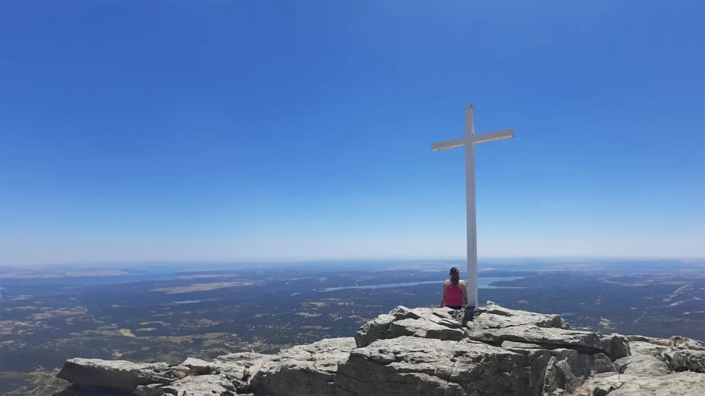 Mulher sentada no Pico de Abantos, perto de uma grande cruz branca, apreciando a vista panorâmica das paisagens vastas e montanhosas ao redor de Madrid em um dia claro e ensolarado.