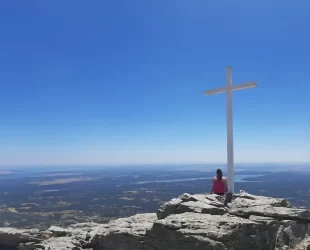 Mulher sentada no Pico de Abantos, perto de uma grande cruz branca, apreciando a vista panorâmica das paisagens vastas e montanhosas ao redor de Madrid em um dia claro e ensolarado.