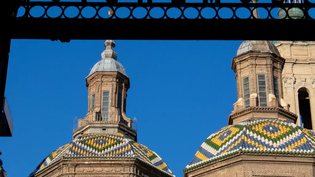 Detalhe das cúpulas da Basílica del Pilar em Zaragoza, Espanha, com telhas coloridas em padrões geométricos, destacando a arquitetura barroca sob um céu azul claro.