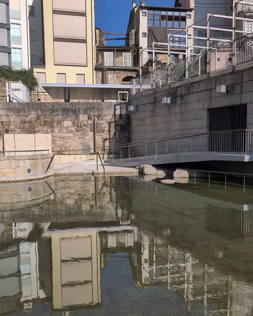 Piscina termal ao ar livre no centro de Ourense, cercada por edifícios e paredes de pedra.