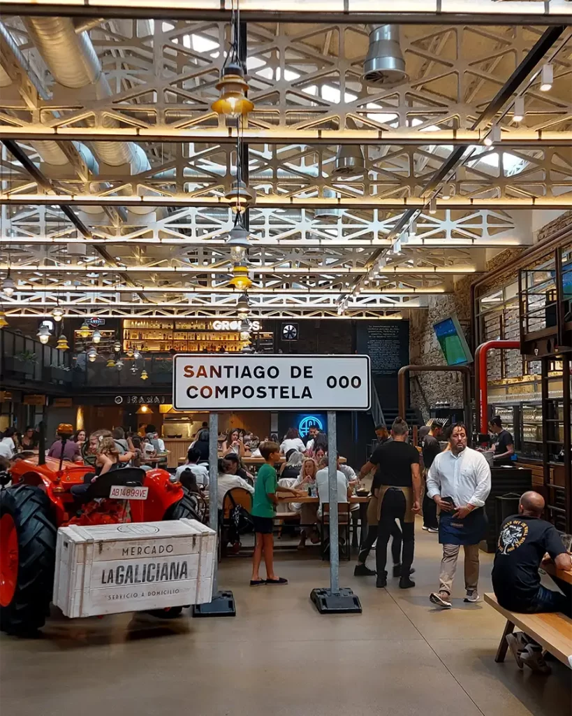 Interior movimentado do Mercado La Galiciana em Santiago de Compostela, com mesas de refeição compartilhadas, um trator vermelho decorativo e uma placa indicando 'Santiago de Compostela'