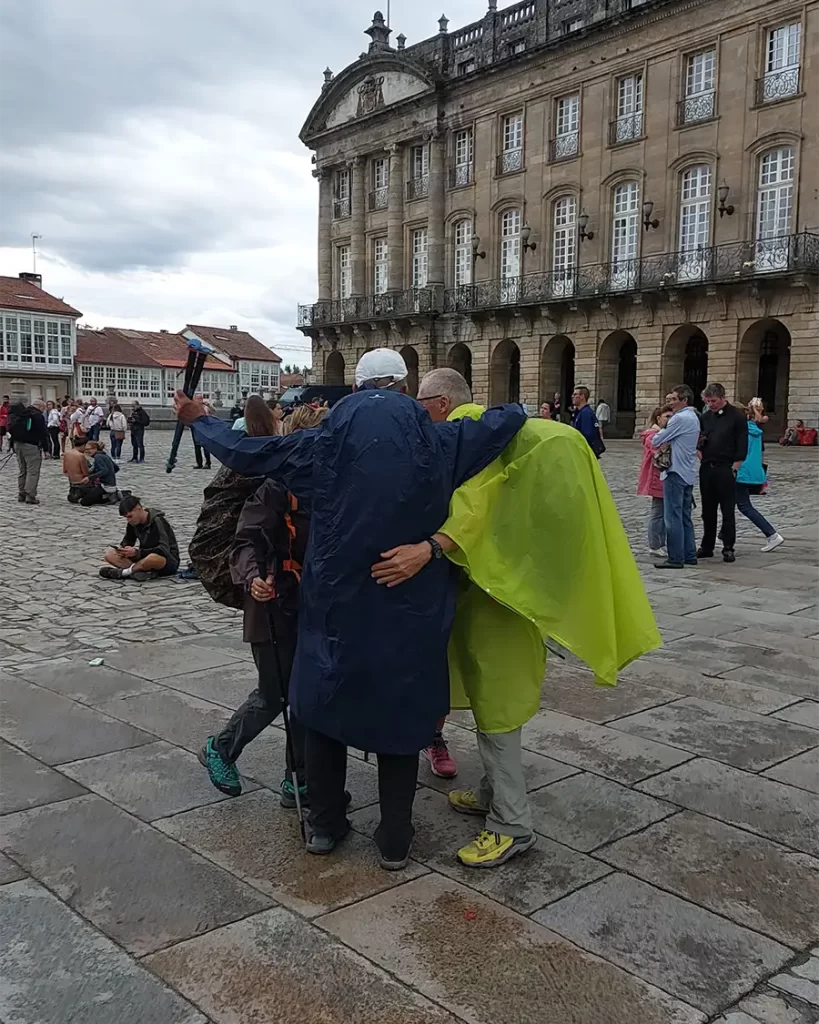 Grupo de peregrinos abraçados na Praça do Obradoiro, com o Palácio de Raxoi ao fundo.