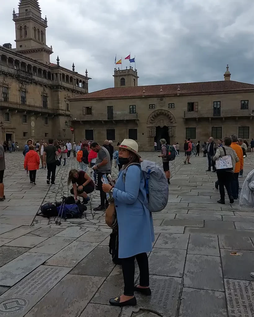 Peregrina com mochila e cajado de caminhada na Praça do Obradoiro, em frente ao Parador dos Reis Católicos, observando a movimentação ao seu redor.