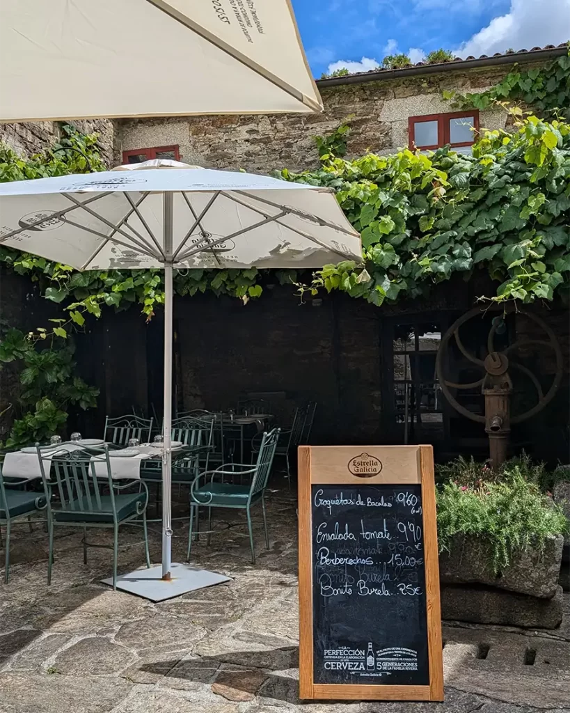 Área externa do Restaurante Sendeiro em Santiago de Compostela, com mesas ao ar livre sob guarda-sóis, cercadas por plantas e uma lousa com o menu do dia.