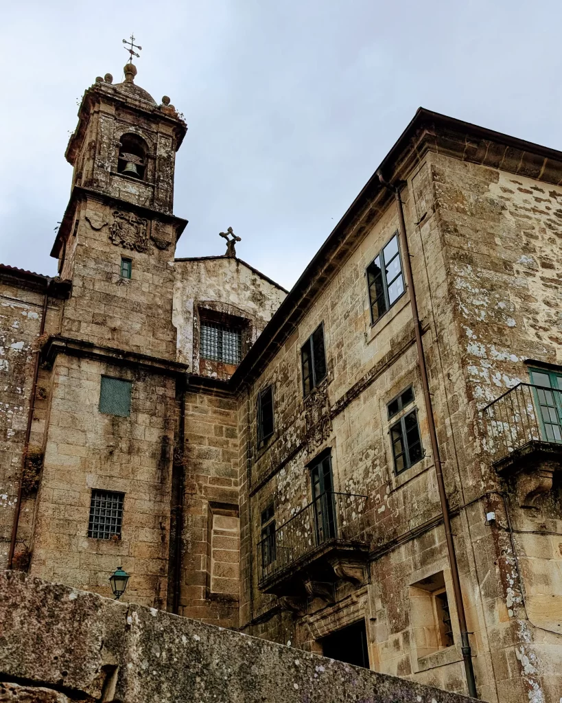 Mosteiro de Belvís visto de baixo, com suas paredes de pedra desgastadas e torre com campanário, localizado próximo ao Parque de Belvís em Santiago de Compostela.