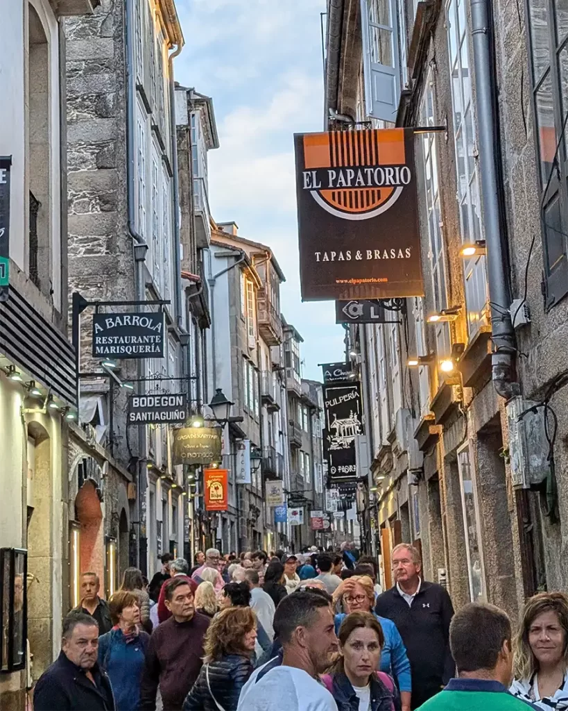 Rua de Santiago de Compostela com vários bares e restaurantes iluminados à noite, cheia de pessoas aproveitando o final do dia.