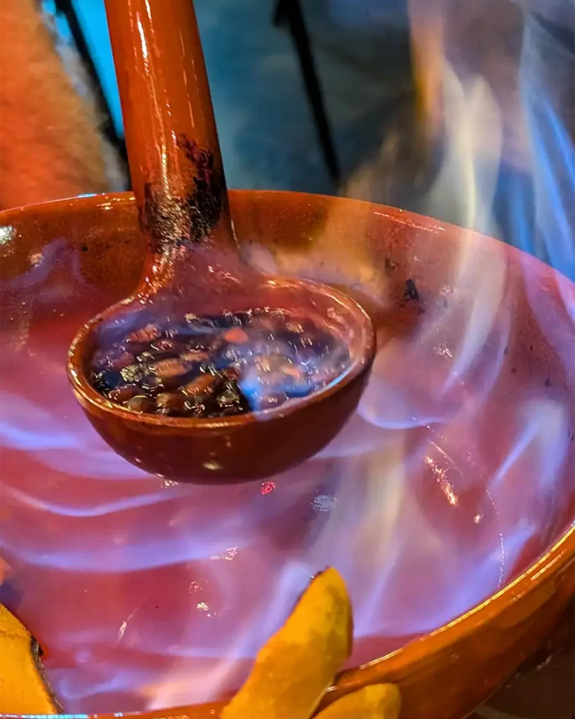 Preparação da Queimada Galega em um recipiente de barro, com a bebida em chamas enquanto é mexida com uma concha, durante o ritual tradicional em Santiago de Compostela.