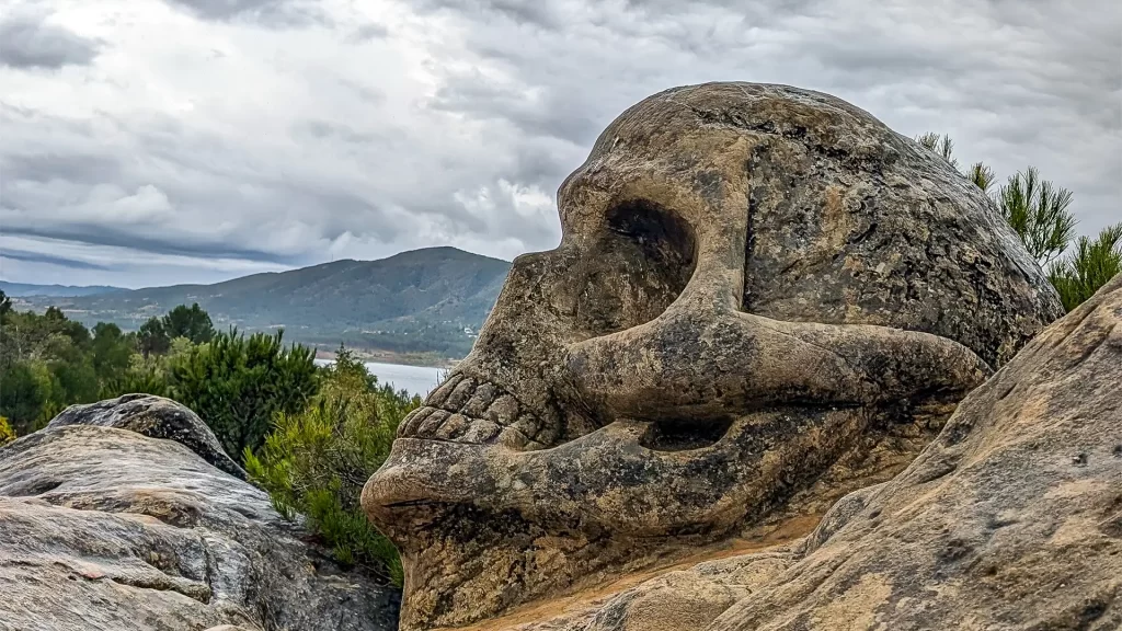 Escultura em forma de crânio esculpida em pedra na Rota das Caras, em Buendía, Cuenca, com montanhas e vegetação ao fundo sob um céu nublado.