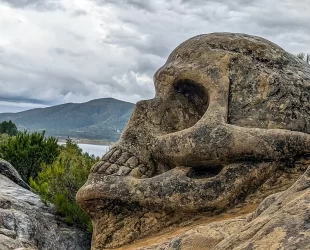 Escultura em forma de crânio esculpida em pedra na Rota das Caras, em Buendía, Cuenca, com montanhas e vegetação ao fundo sob um céu nublado.