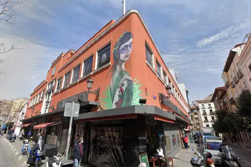 Fachada do Mercado de Antón Martín, em Madrid, com paredes vermelhas decoradas por um grande mural artístico. A entrada principal possui portas de vidro que permitem vislumbrar o interior vibrante do mercado.
