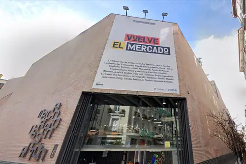 Fachada do Mercado de San Antón, em Madrid, com design moderno e minimalista. Um grande cartaz anuncia "Vuelve el Mercado," destacando a revitalização do espaço. A entrada de vidro permite vislumbrar o interior vibrante do mercado gastronômico.