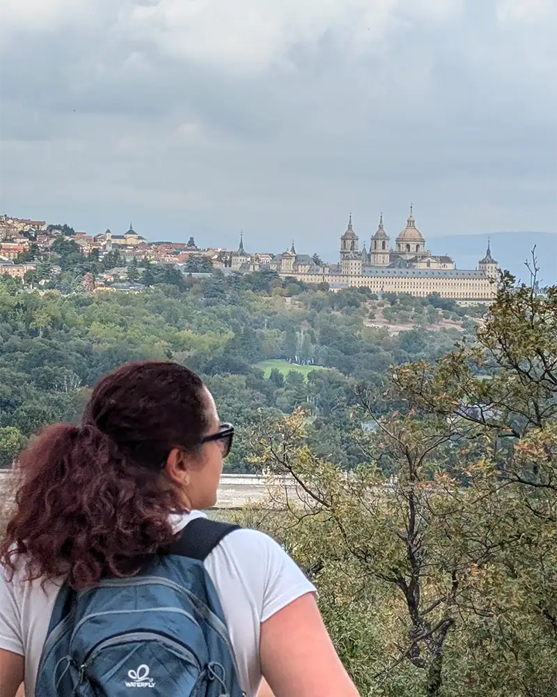 Eu observando o Mosteiro de El Escorial ao longe, de uma trilha cercada por árvores.