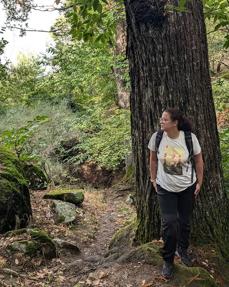 Eu encostada em uma árvore robusta no bosque, rodeada por vegetação densa e trilhas.