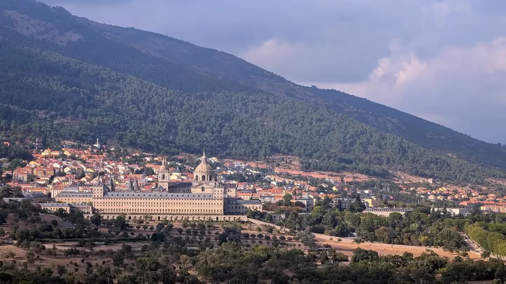 Mosteiro de El Escorial cercado por casas do vilarejo e colinas verdes sob céu nublado.