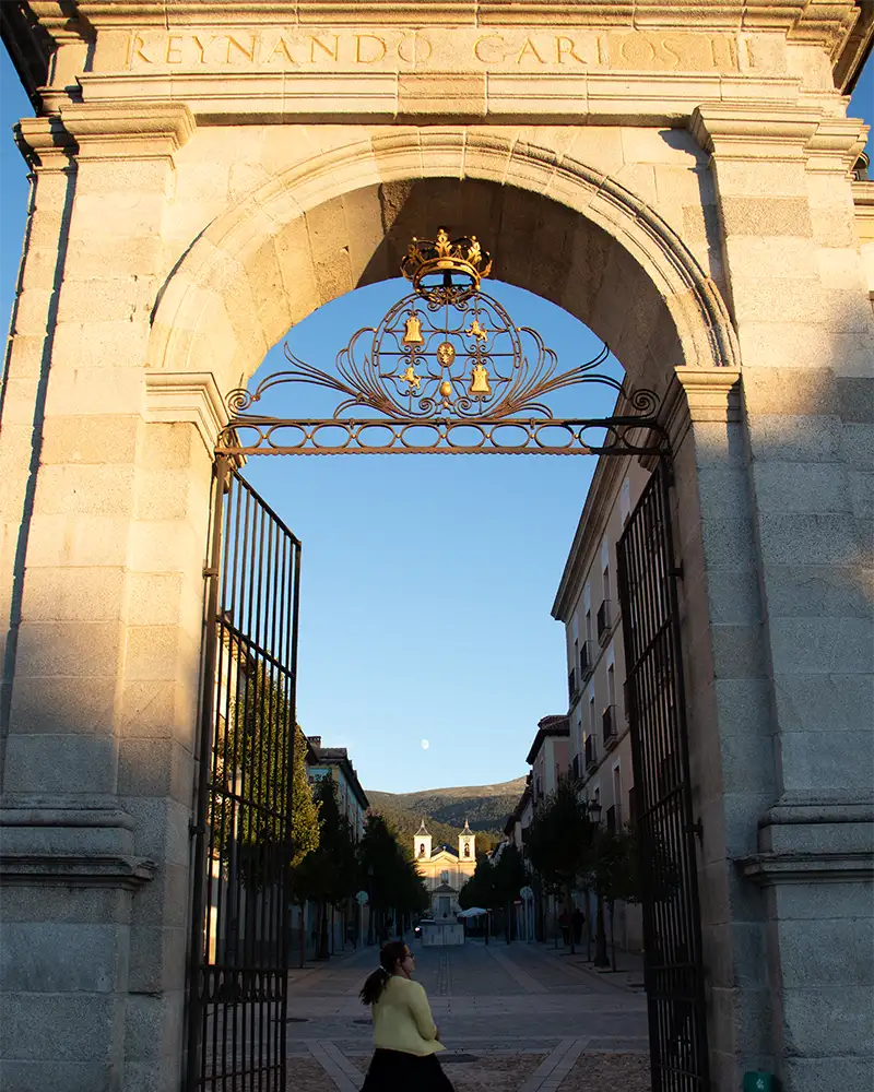 Arco de entrada em granito marcado com 'Reynando Carlos III', com uma pessoa caminhando ao centro e montanhas ao fundo em San Ildefonso.