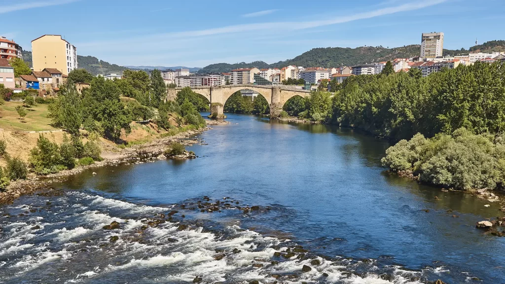 Vista da Ponte Romana sobre o Rio Minho em Ourense, Galícia, cercada por vegetação e prédios ao fundo em um dia ensolarado.