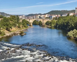 Vista da Ponte Romana sobre o Rio Minho em Ourense, Galícia, cercada por vegetação e prédios ao fundo em um dia ensolarado.