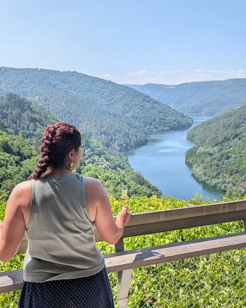 Uma mulher segurando uma taça de vinho branco enquanto aprecia a vista do rio Sil em um mirante cercado por vinhedos.