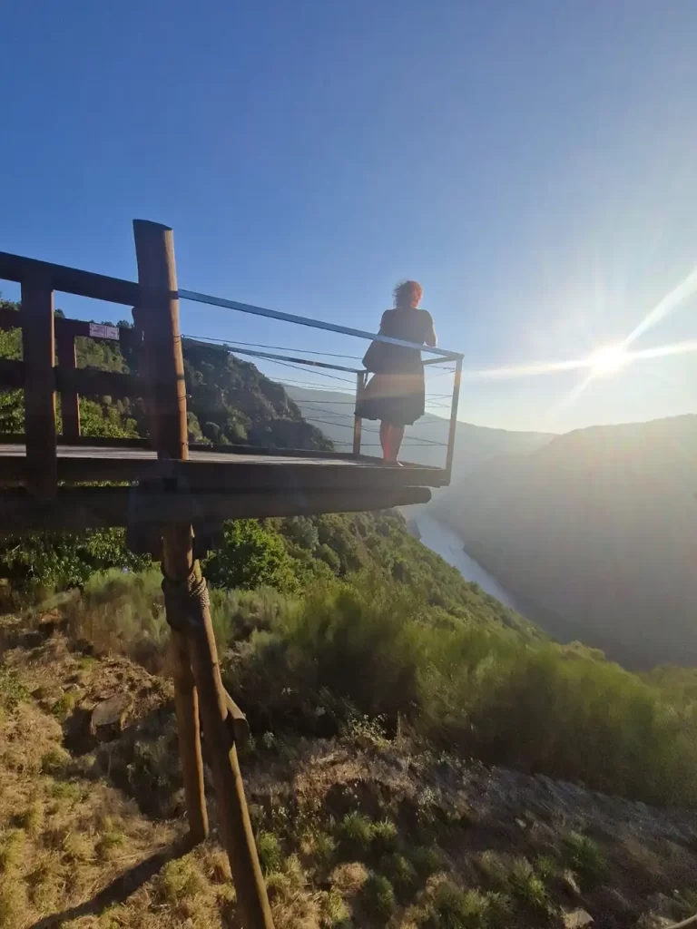 Uma pessoa em um mirante de madeira admirando o rio Sil e as colinas verdes sob um céu ensolarado.