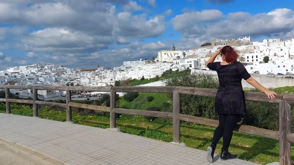 Vista panorâmica de Vejer de la Frontera, com suas casas brancas características sob um céu parcialmente nublado. Em primeiro plano, a autora do artigo aprecia a paisagem de um mirante.