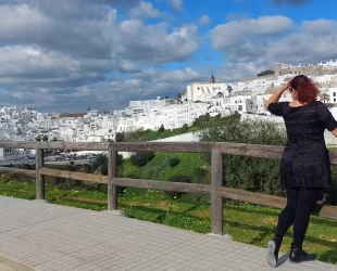 Vista panorâmica de Vejer de la Frontera, com suas casas brancas características sob um céu parcialmente nublado. Em primeiro plano, a autora do artigo aprecia a paisagem de um mirante.