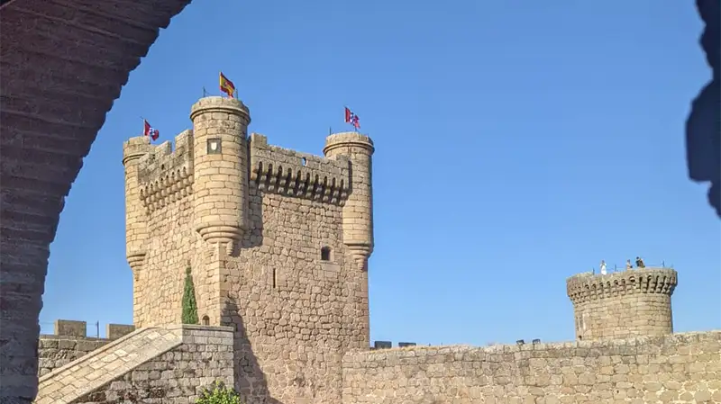 Torre principal do Parador de Oropesa, um castelo de pedra com bandeiras no topo, visto através de um arco. O céu está claro e azul.