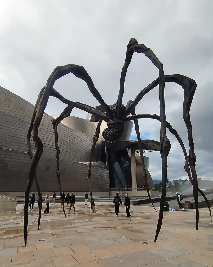 Escultura de aranha Maman de Louise Bourgeois proxima ao rio, com visitantes e predios ao fundo em Bilbao