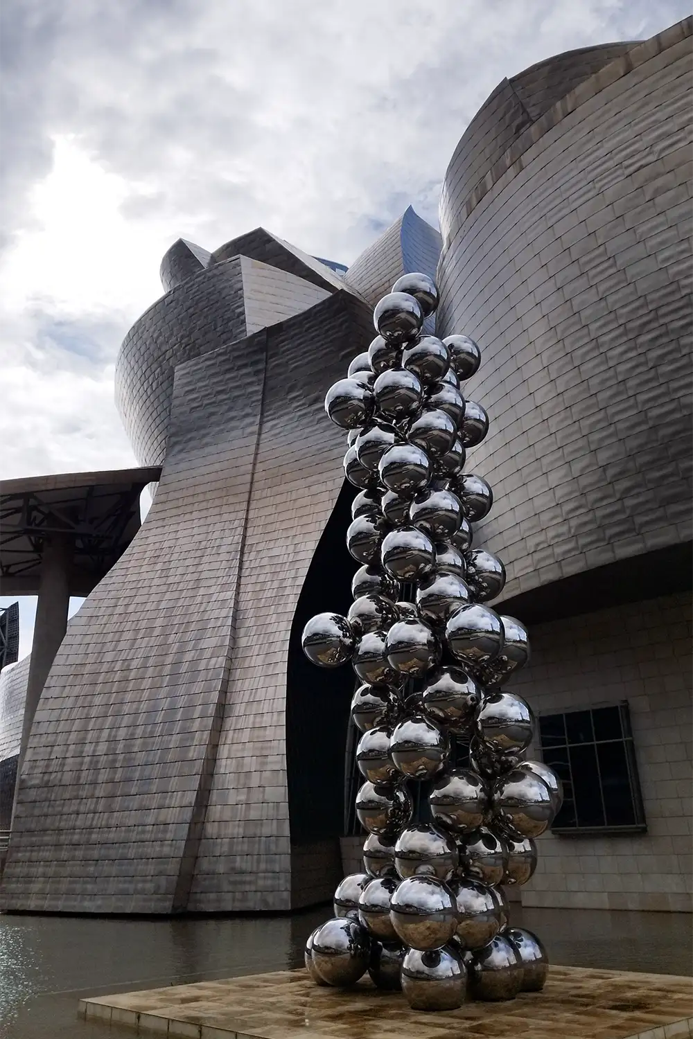 Fachada do Museu Guggenheim Bilbao com escultura de esferas metálicas refletoras à beira do rio
