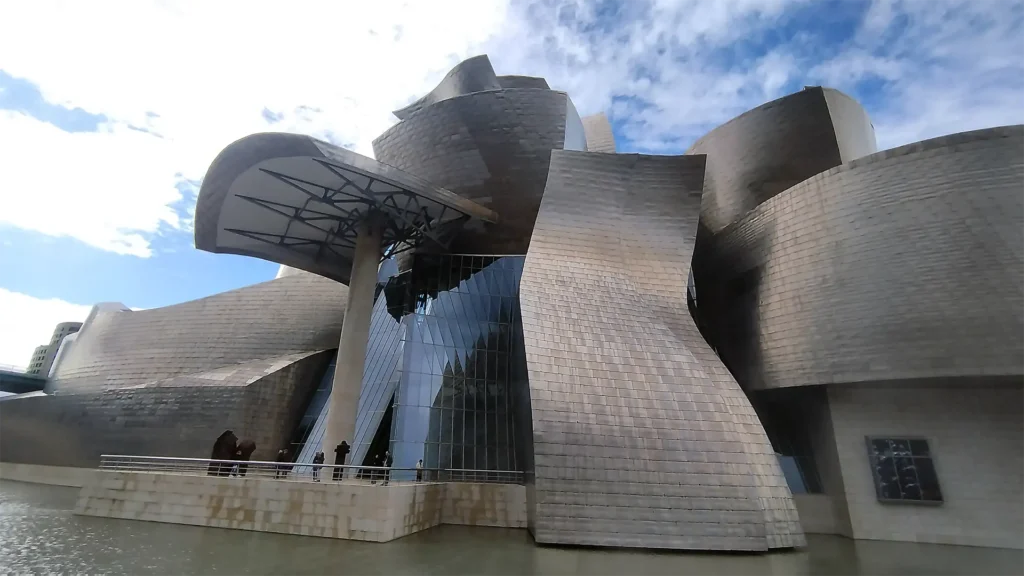 Vista lateral da fachada do Museu Guggenheim Bilbao, com volumes metálicos curvos e assimétricos que refletem a luz do sol.
