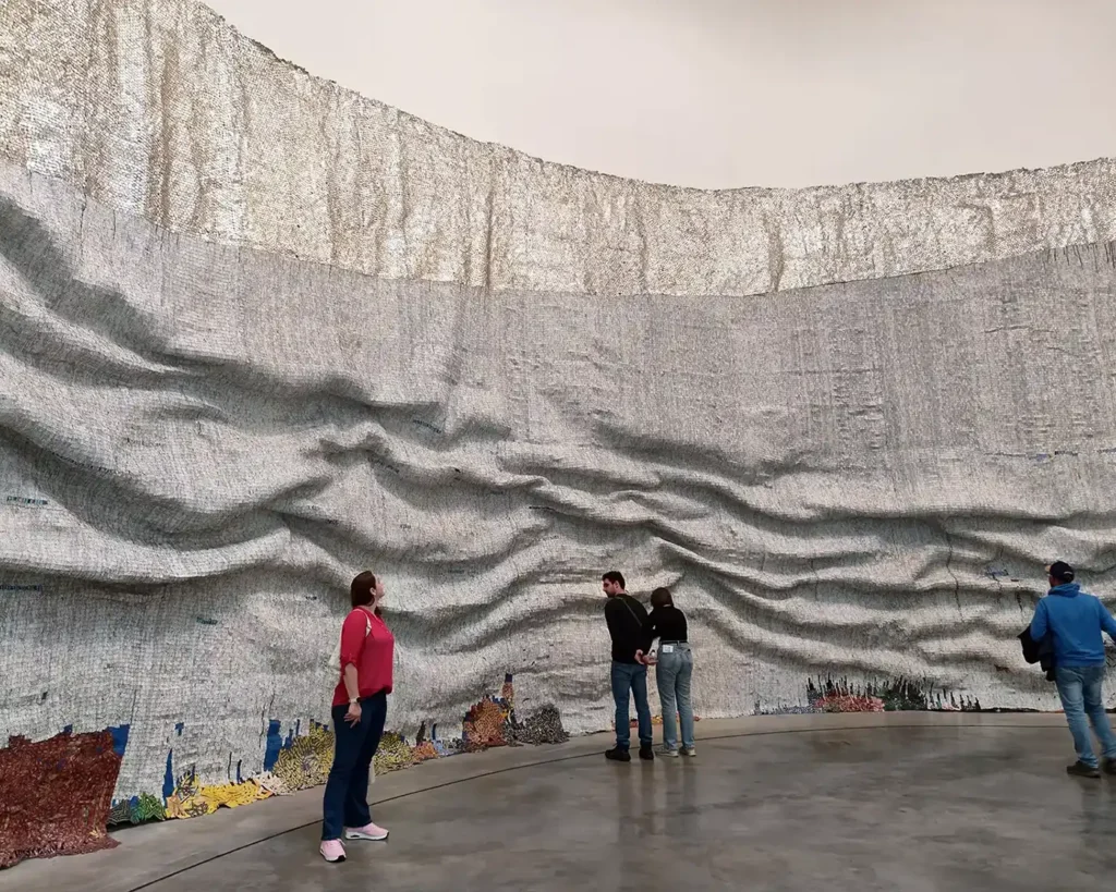 Visitantes observando a instalacao Rising Sea de El Anatsui no Guggenheim Bilbao, feita com tampas de aluminio