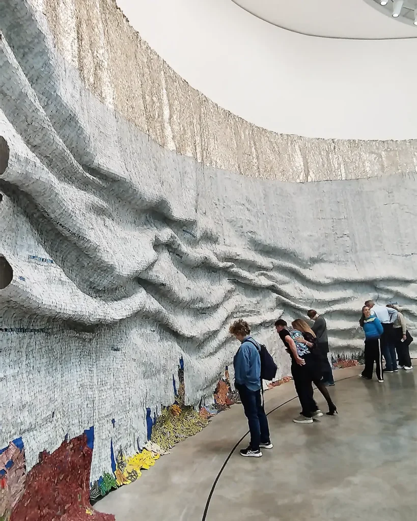 Detalhe da obra Rising Sea de El Anatsui com visitantes em frente ao painel metalico ondulado