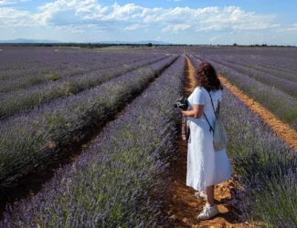 Mulher de branco com câmera, de costas, andando por vasto campo de lavanda roxa sob céu azul com nuvens em Brihuega