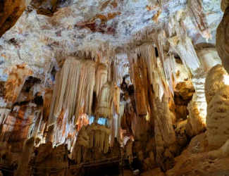 Interior da Cuevas del Águila, mostrando um grande salão com estalactites e estalagmites de diferentes tamanhos e formas, iluminadas para destacar as formações minerais.