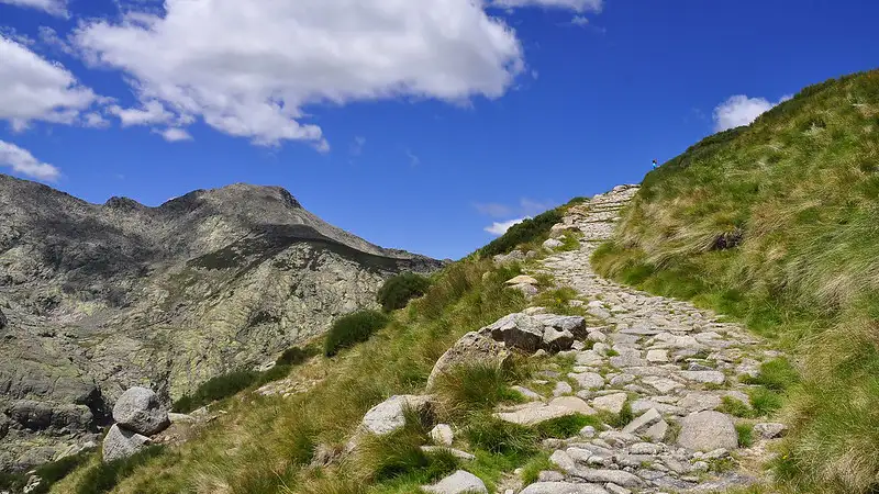 Caminho de pedras da Calçada Romana subindo uma montanha com grama, com os picos rochosos da Serra de Gredos ao fundo.